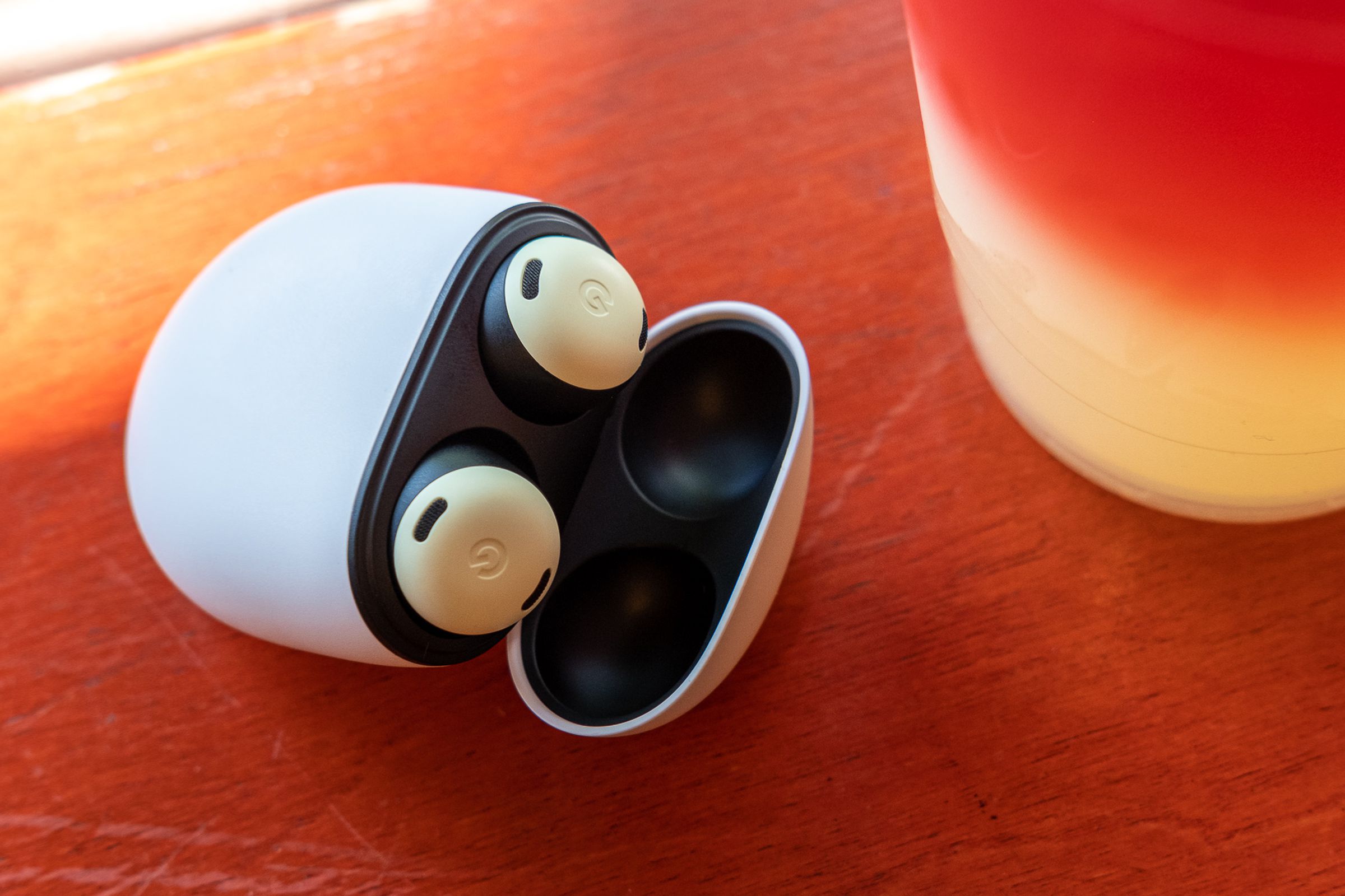 The yellow Pixel Buds Pro wireless earbuds sitting in their charging case with the lid open, resting on an orange table beside a plastic cup of colorful lemonade.