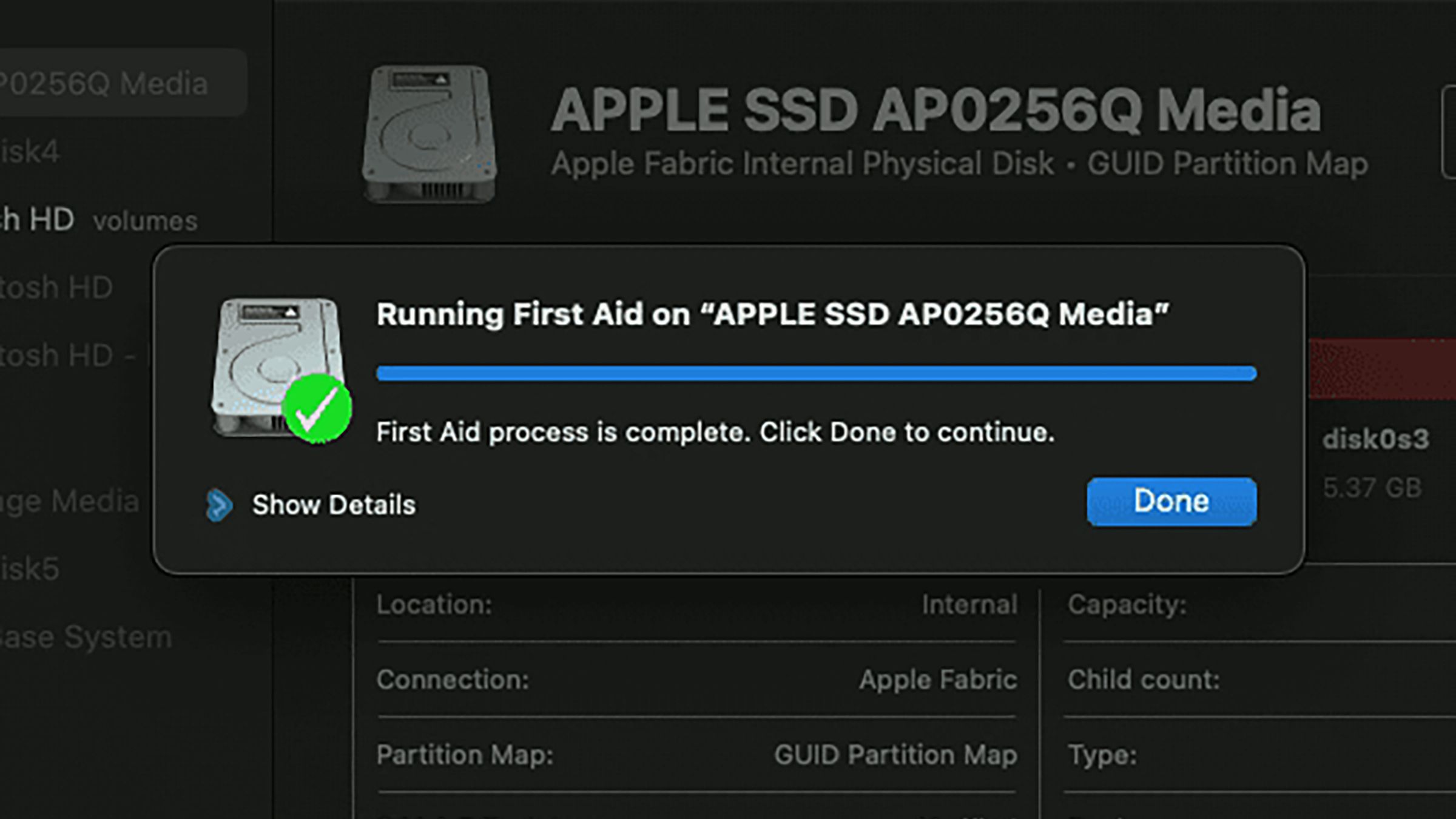 The Mac Disk Utility tool showing info about the Mac in background, and a pop-up with a photo of a hard drive, a long blue line, and a caption “Running First Aid on “Apple SSD AP0256Q Media”