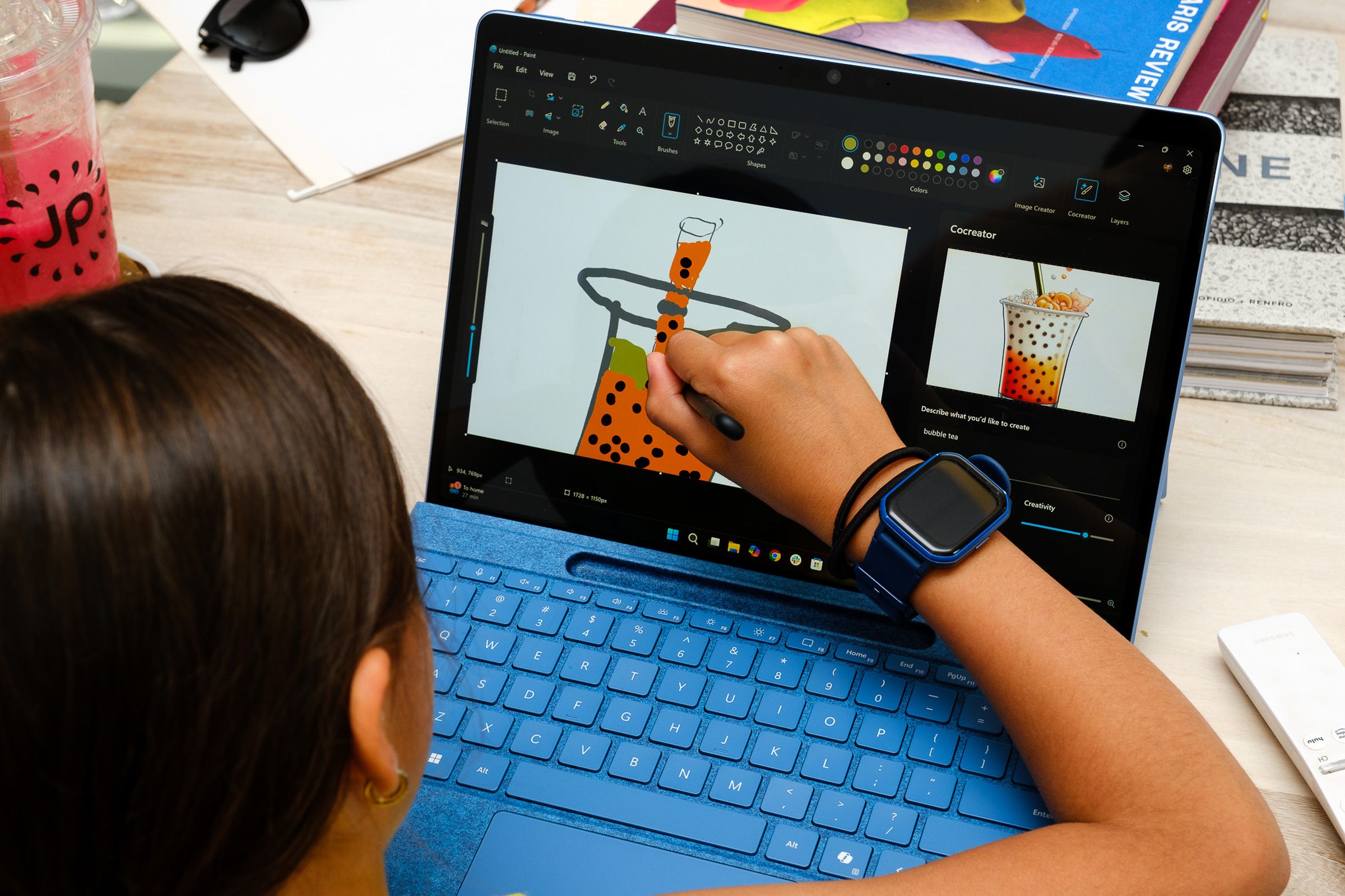 A photo of a young person using a stylus to draw a picture of a bubble tea in MS Paint, on the Surface Pro. Paint Cocreator has rendered a slightly fancier bubble tea image on the right side of the screen. 