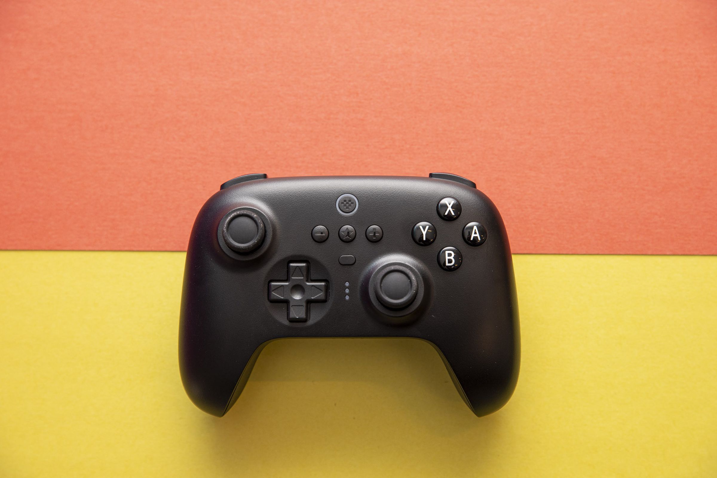 Top-down shot of 8BitDo Ultimate Controller on an orange and yellow background. The controller has an asymmetric joystick design, like an Xbox or Switch Pro controller, and it’s black, with white legends on the A B X Y buttons.