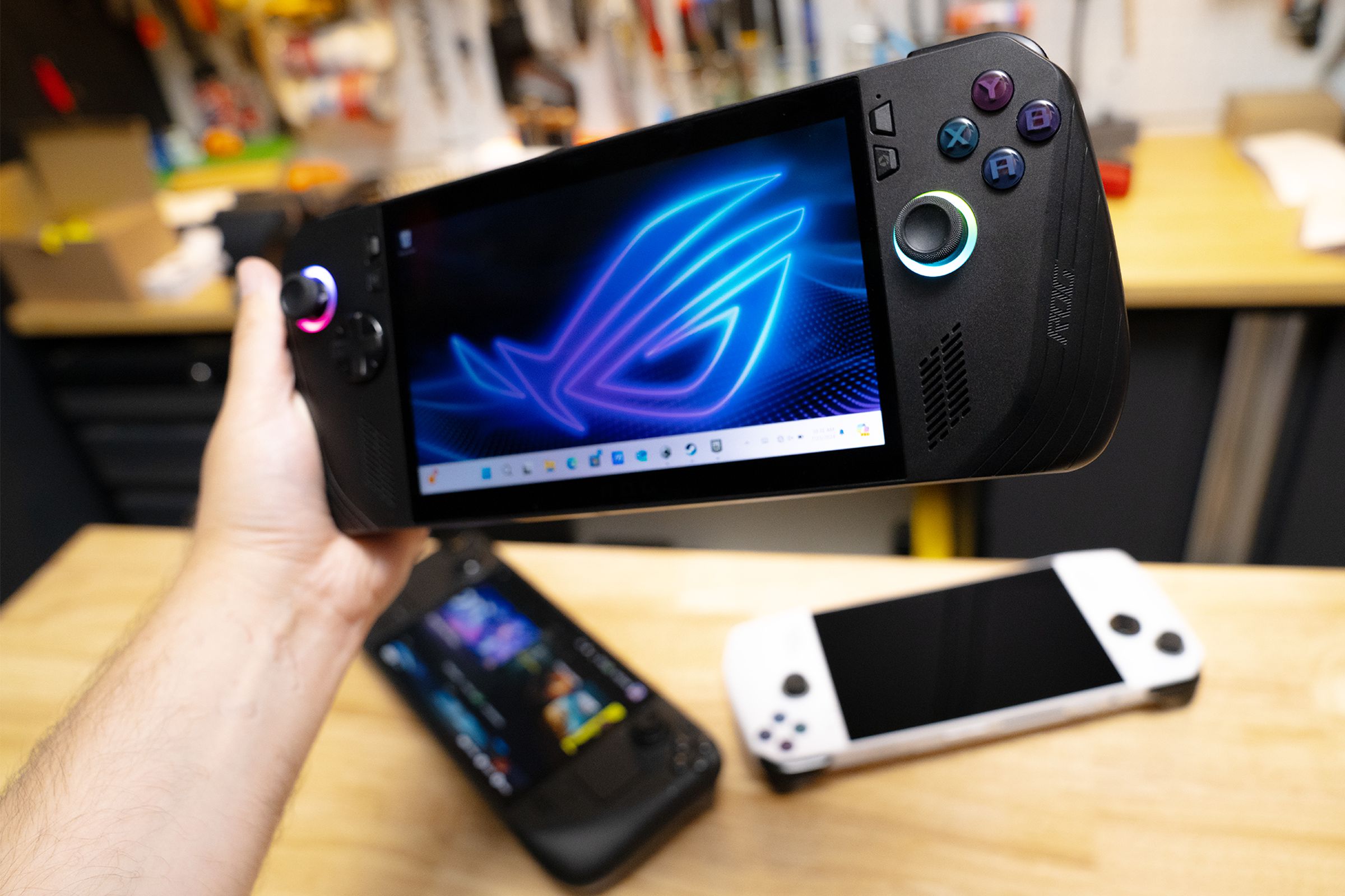 The new black ROG Ally X handheld gaming PC is held above a wooden surface containing an original white ROG Ally and the Steam Deck OLED.