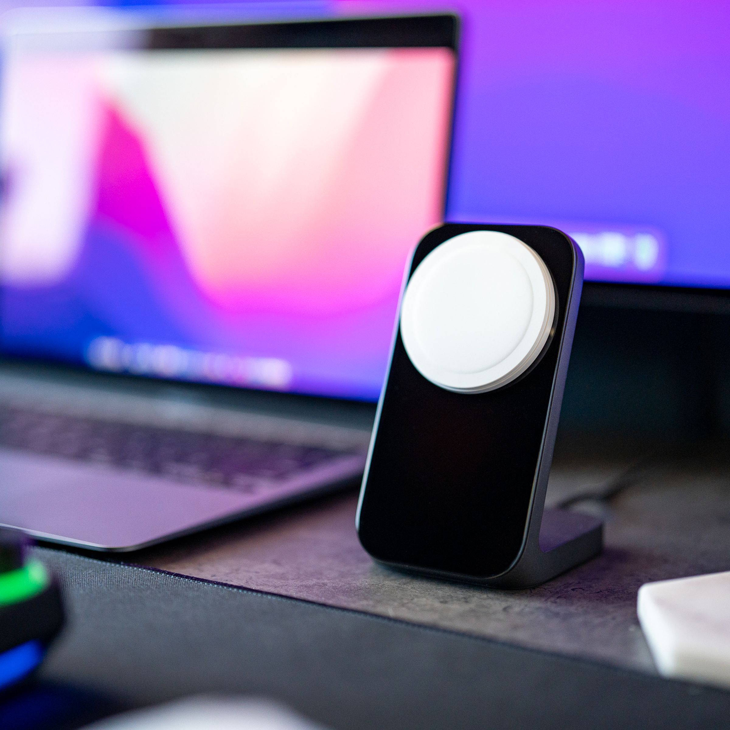 The Nomad Stand One MagSafe charger in black, sitting on a gray desk beside a MacBook Air, keyboard, and monitor.