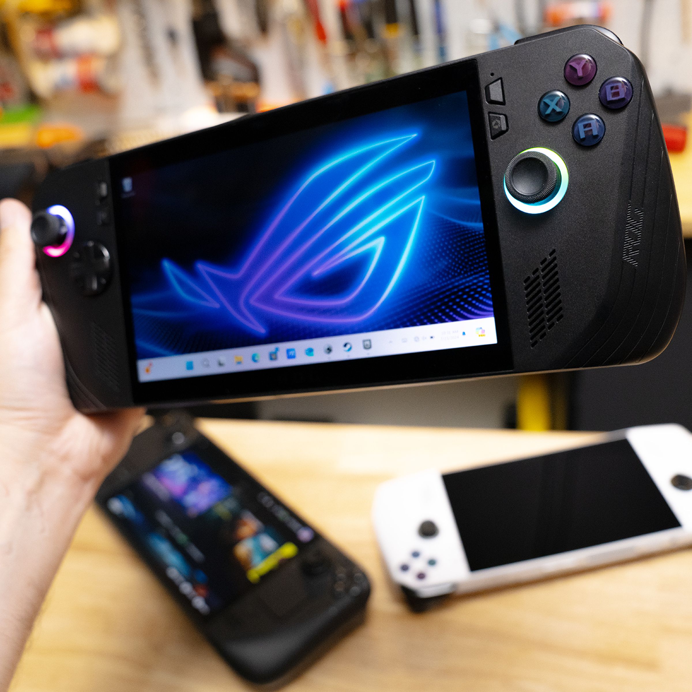 The new black ROG Ally X handheld gaming PC is held above a wooden surface containing an original white ROG Ally and the Steam Deck OLED.