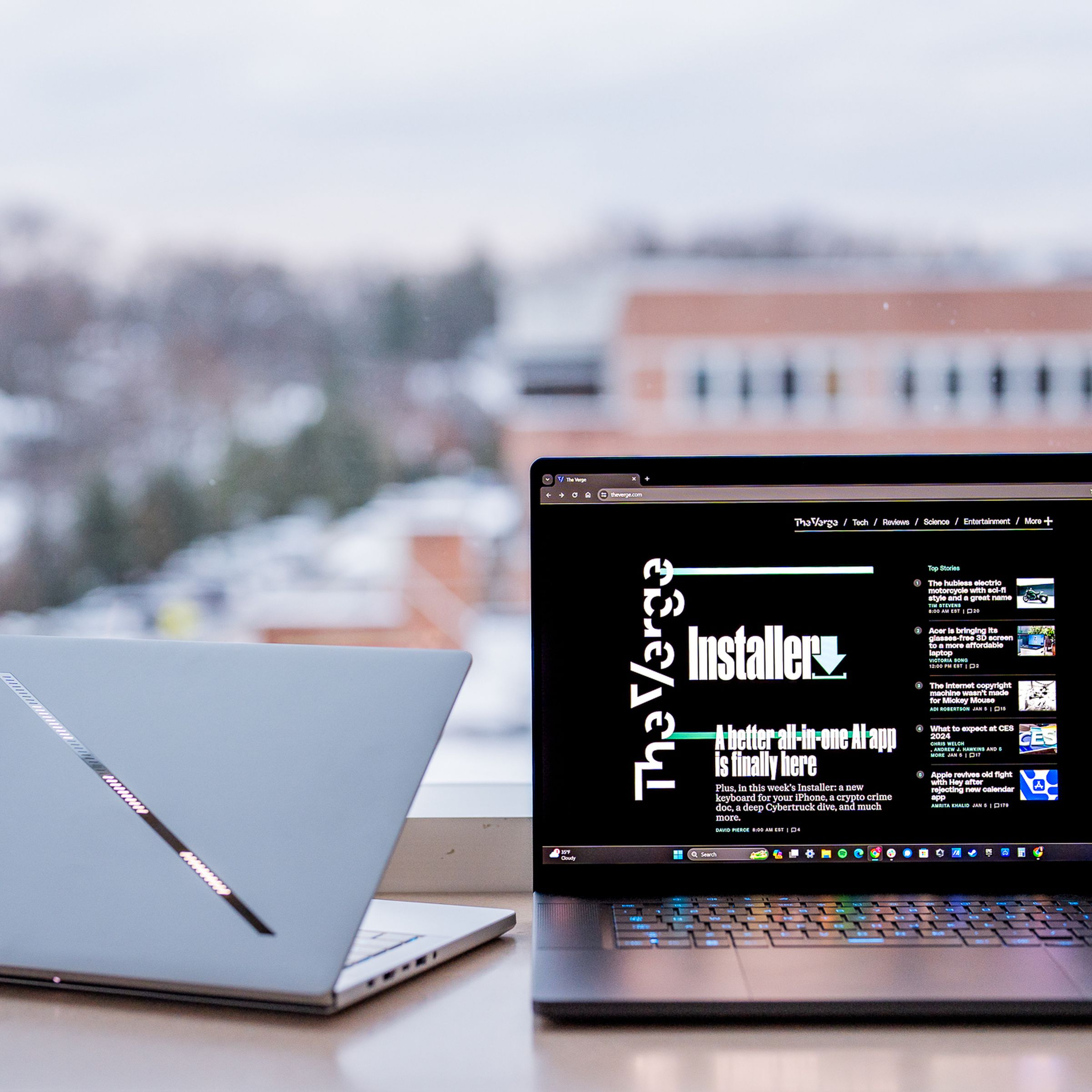 A sleek silver gaming laptop, on left, facing away from camera. A shiny slash is the sole decoration on the lid, running from top left to bottom right corners. On the right is a second, larger gaming laptop, screen open, with the Verge homepage displayed. 