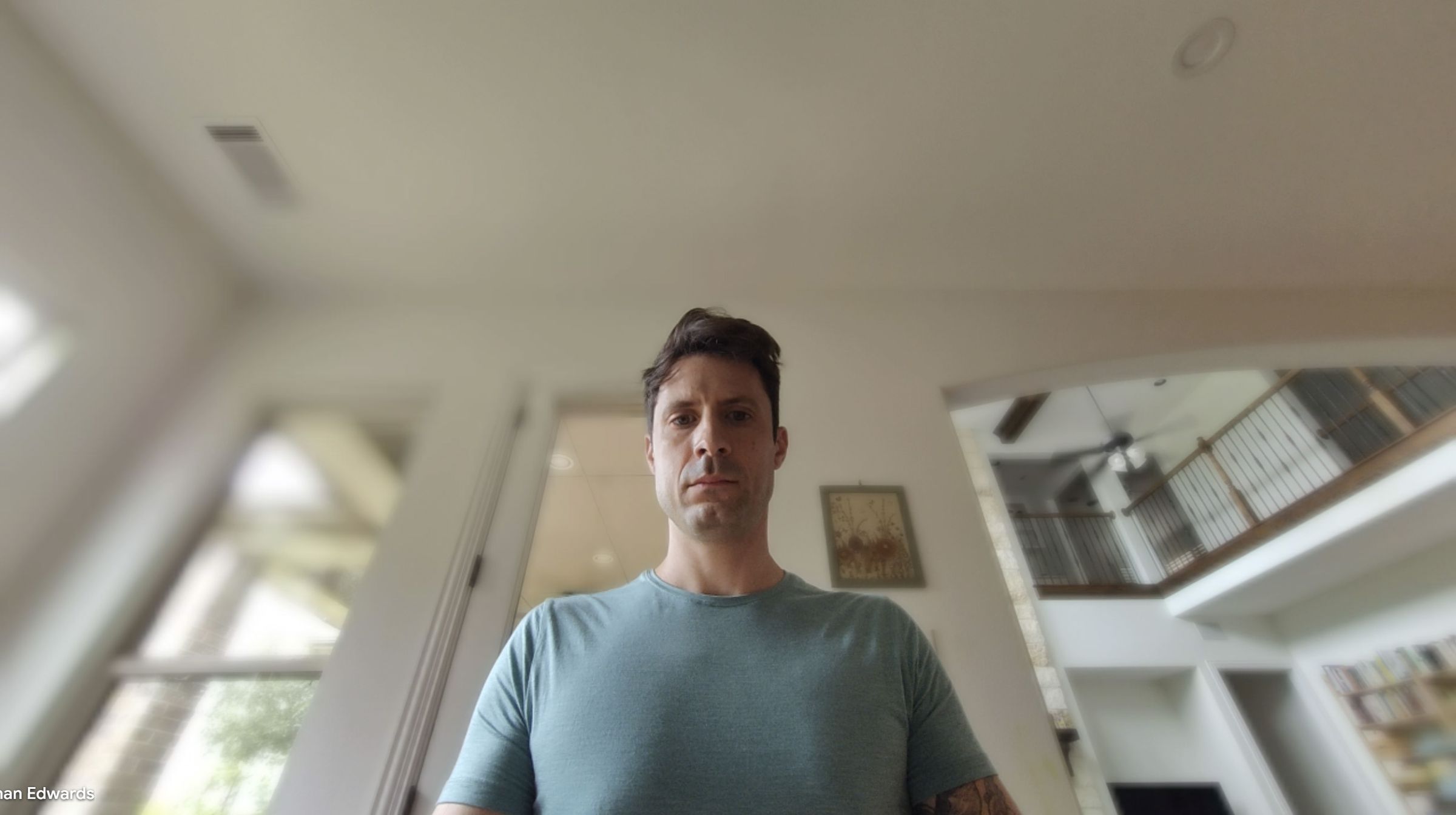 A webcam shot of a man in a teal shirt. The upward angle and wide lens make the background look vast.