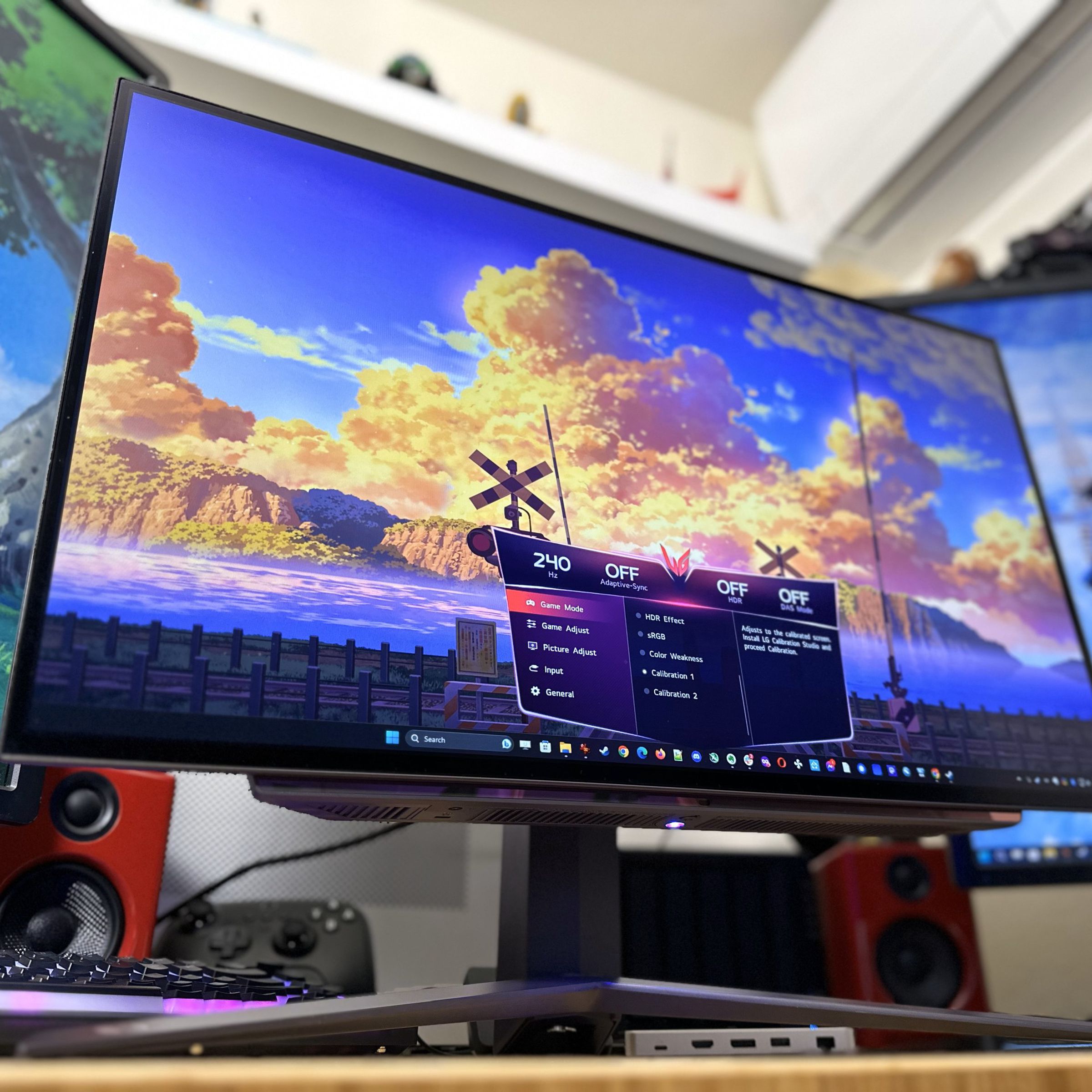 A bright, colorful OLED monitor with a big V-shaped stand underneath, flanked by two other monitors in portrait mode. The main monitor has a colorful sky on screen with big orange sunset-lit clouds and an on-screen display showing it’s running at 240Hz.