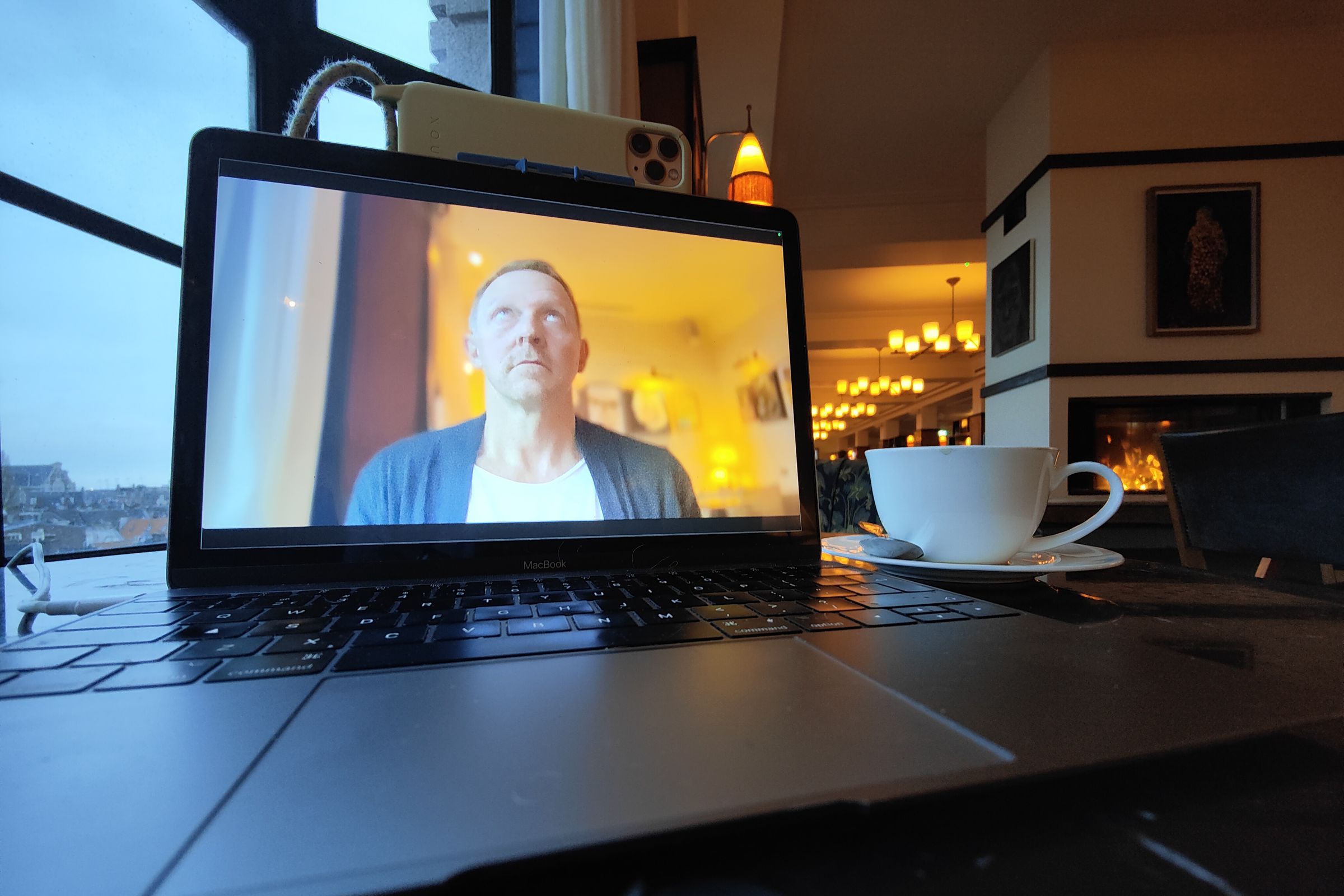 Man’s face is being recorded on a MacBook display using an iPhone mounted to the top of the MacBook. The environment is cozy and dimly lit.