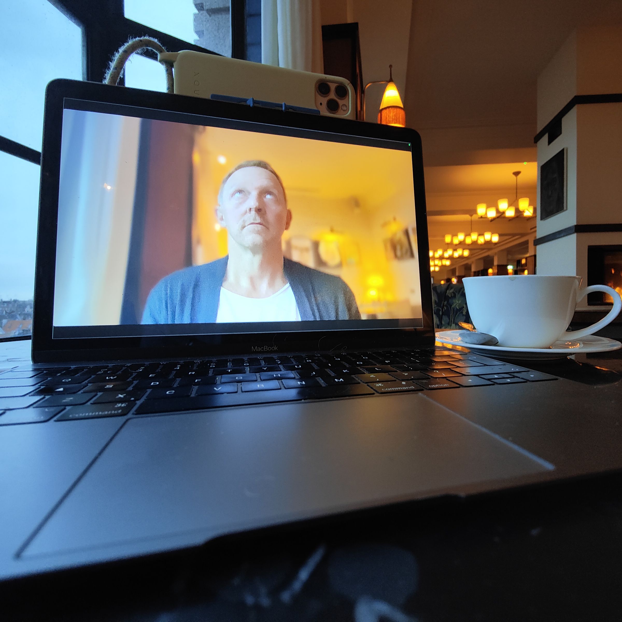 Man’s face is being recorded on a MacBook display using an iPhone mounted to the top of the MacBook. The environment is cozy and dimly lit.