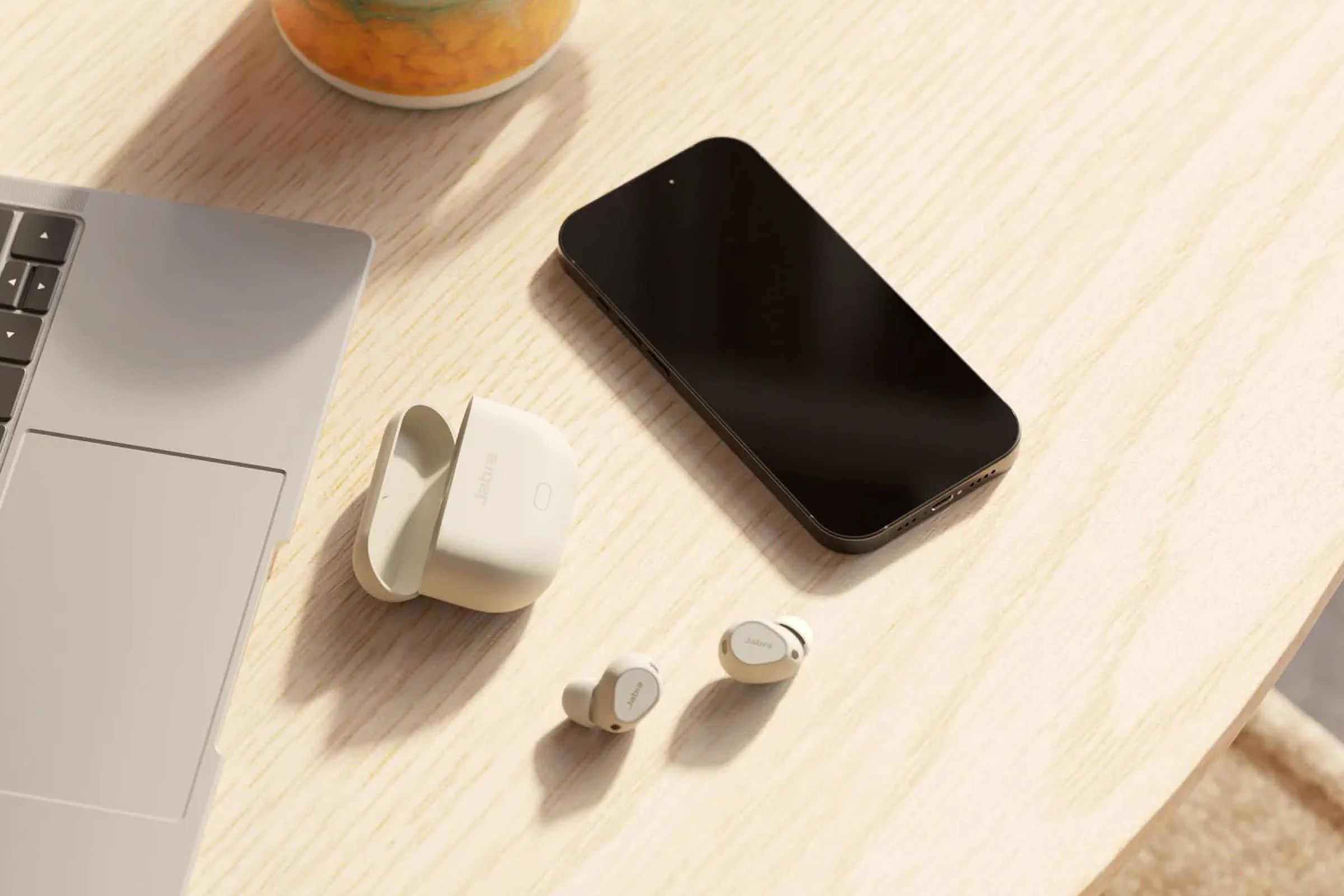 The Jabra Elite 10 gen 2 wireless earbuds on a table besides a laptop and a smartphone.