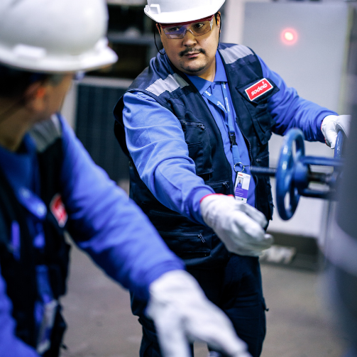 Two men wearing hard hats and white gloves, working on a machine.