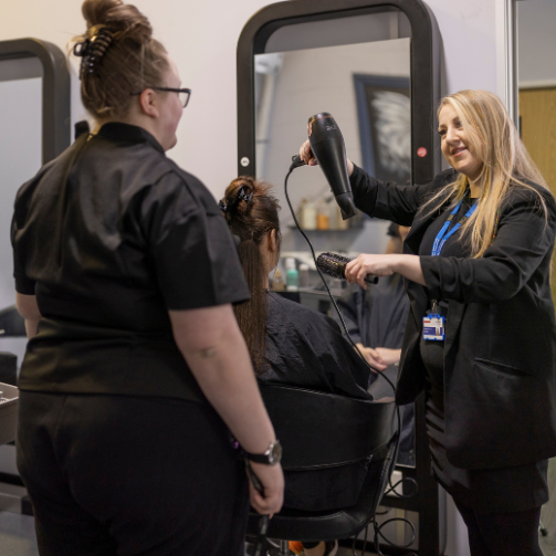 Woman in a full black outfit showing another woman how to style someone’s hair. 