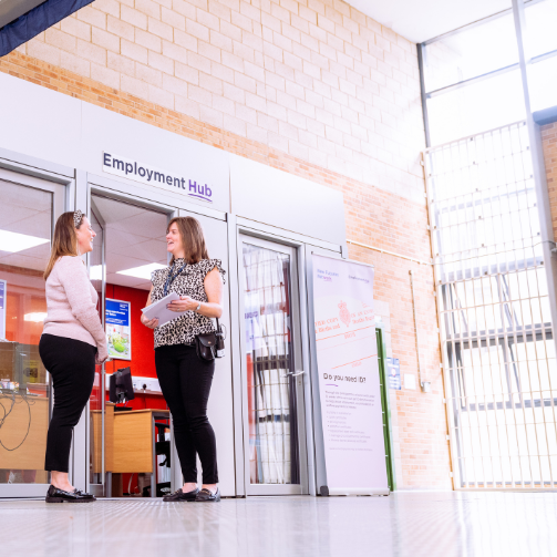 Two women having a conversation in front of an office with a sign that reads Employment Hub.