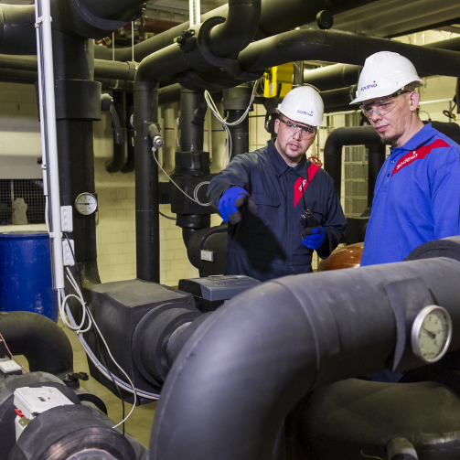 Two men in blue shirts and hard hats working on some pipes.