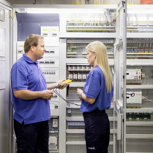 A man and a woman in blue uniforms standing by technical equipment.