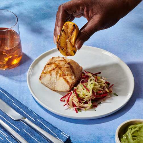 Hand of a person adding a tangy touch to a piece of fish by holding a lemon slice above it.