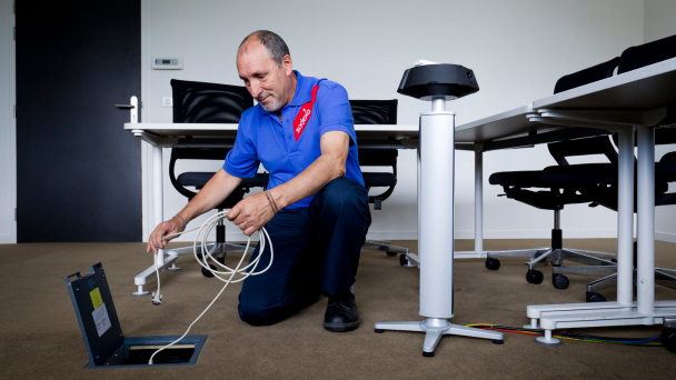 A man kneeling to fix a cable in an office, surrounded by computers and office equipment