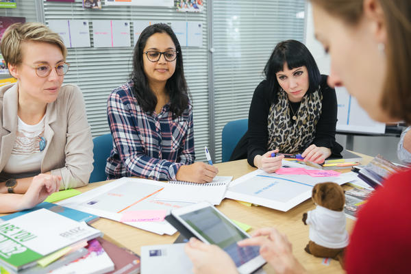 Three women sat around a table in a workshop. There are papers and booklets on the table and they are smiling. 