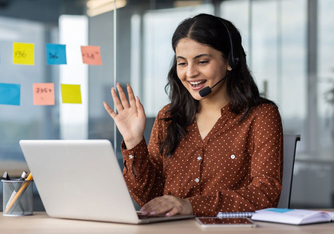 Mercado Pago: imagem de profissional de atendimento ao cliente realizando um processo de prós-venda. Na imagem, é possível ver a profissional sentada à mesa de trabalho em frente ao notebook com um headset, com uma das mãos levantada, indicando que está realizando um atendimento por videochamada. Sobre a mesa, é possível ver também um caderno e canetas.