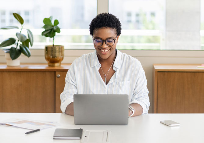 Mercado Pago: Mulher de óculos e vestindo uma camisa branca enquanto pesquisa como precificar um produto dropshipping em um laptop em um escritório moderno e iluminado, com uma planta e itens de escritório ao fundo.