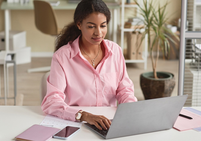 Mercado Pago: empresária de camisa rosa sentada em mesa em seu escritório, com notebook na frente realizado o controle de estoque de sua empresa. Na mesa, também há uma caneca, celular e pastas. 