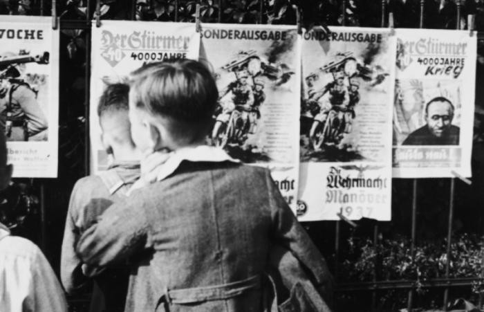 A group of young German boys view "Der Stuermer," "Die Woche," and other propaganda posters that are posted on a fence in Berlin, Germany, 1937.