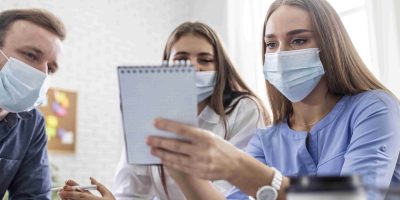 Três estudantes do curso de Enfermagem usando máscaras de proteção participam de uma reunião colaborativa em um ambiente de escritório, analisando anotações em um caderno.