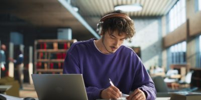 Estudante concentrado em biblioteca, revisando a lista de obras literárias obrigatórias da Fuvest com fones de ouvido e materiais de estudo.