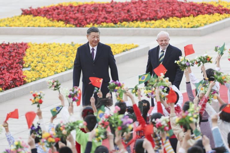 Na imagem, duas figuras masculinas caminham em direção a um grupo de pessoas que seguram flores e bandeiras vermelhas. O fundo é decorado com um grande canteiro de flores coloridas, predominantemente amarelas e vermelhas. As pessoas no grupo estão vestidas com roupas tradicionais e parecem estar celebrando.