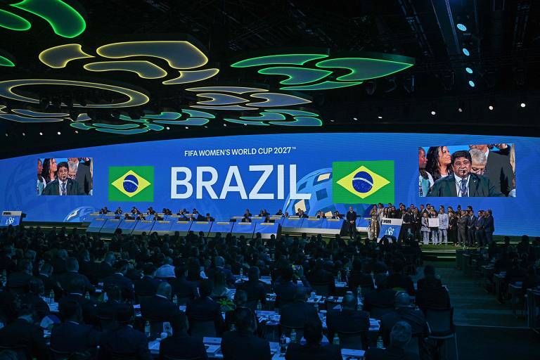 A imagem mostra um evento relacionado à FIFA, onde o Brasil foi anunciado como sede da Copa do Mundo Feminina de 2027. No fundo, há um grande telão exibindo o texto 'FIFA WOMEN'S WORLD CUP 2027' e a palavra 'BRAZIL', acompanhada da bandeira do Brasil. À frente, uma mesa com vários participantes e um orador em destaque, enquanto a plateia é composta por pessoas em trajes formais.
