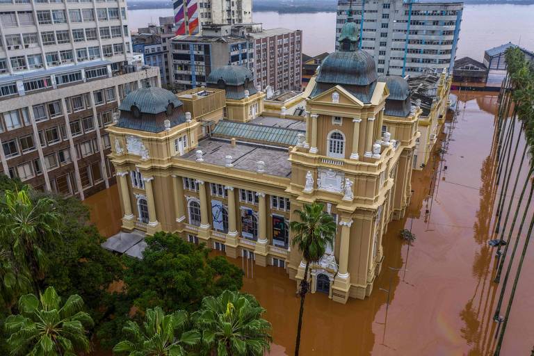 A imagem mostra um edifício histórico, com uma arquitetura clássica, parcialmente submerso em água de cor marrom, resultado de uma inundação. O prédio possui cúpulas e colunas, e está cercado por árvores e palmeiras. Ao fundo, há prédios modernos e uma vista do rio. A água cobre a área ao redor, incluindo ruas e postes de iluminação.