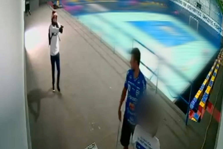 Na foto, um homem de camisa branca aponta uma arma para outro homem de camisa azul. Ao fundo, uma cancha de futsal