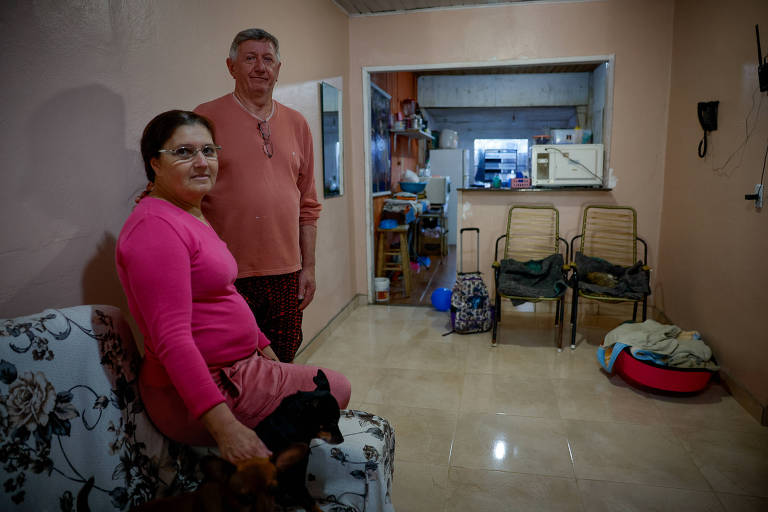 A imagem mostra um casal em uma sala de estar. A mulher, vestindo uma blusa rosa e uma saia, está sentada em um sofá com estampa floral, segurando um cachorro pequeno. O homem, usando uma camiseta rosa claro e calças, está em pé ao lado dela. Ao fundo, há uma cozinha visível, com móveis e utensílios. O ambiente é iluminado e possui paredes de cor clara.