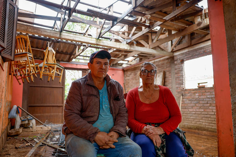 A imagem mostra um homem e uma mulher sentados em um ambiente com o teto danificado. O homem usa uma jaqueta marrom e uma camiseta azul, enquanto a mulher está vestida com uma blusa vermelha e óculos. Ao fundo, há cadeiras empilhadas e paredes de tijolos expostos, indicando um espaço que precisa de reparos.