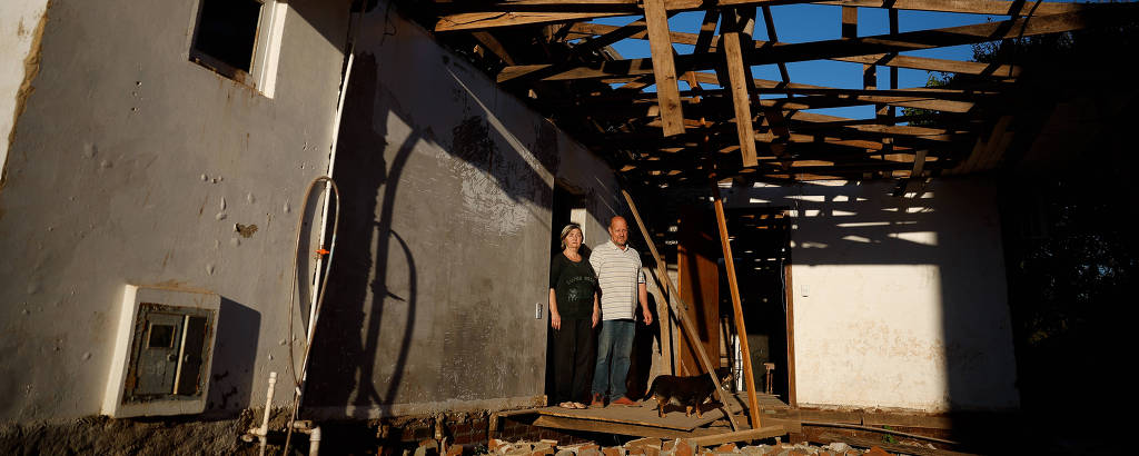 A imagem mostra duas pessoas em pé dentro de uma casa com o telhado danificado. O teto está parcialmente desmoronado, com vigas de madeira expostas e uma parte do telhado faltando. As paredes da casa são de cor clara e há uma janela visível. O chão está coberto por pedras e detritos. A luz do sol ilumina a cena, criando um contraste entre as sombras e as áreas iluminadas.