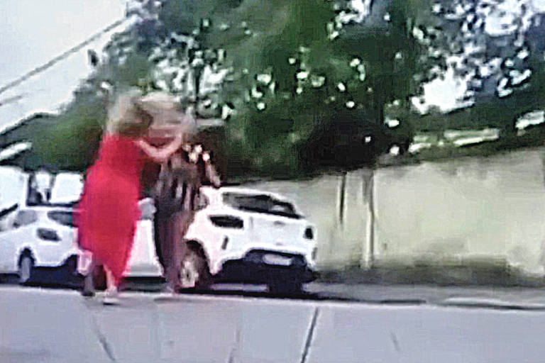 Na imagem, uma mulher loira de vestido vermelho é agredida por uma outra mulher de roupa escura no meio de uma rua.