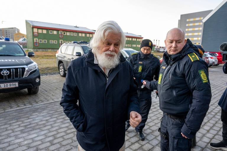 A imagem mostra um homem de barba branca e cabelo longo, vestido com um casaco escuro, caminhando em direção a um grupo de policiais. Ele parece estar sendo acompanhado por dois policiais, um deles com um uniforme escuro e o outro com um uniforme claro. Ao fundo, há veículos policiais e um edifício com janelas.