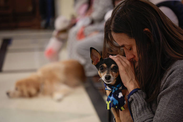 Mulher está com a cabeça abaixada, encostrada na de seu cachorrinho, que olha em direção à câmera