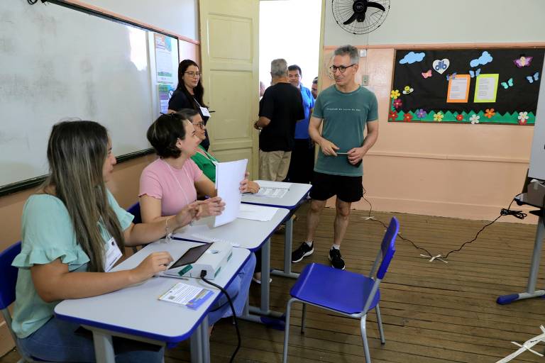 A imagem mostra uma sala de aula com várias pessoas. À esquerda, três mulheres estão sentadas em mesas, uma delas usando uma blusa verde, outra uma blusa rosa e a terceira uma blusa verde. Elas estão interagindo e segurando papéis. Um homem de camiseta verde e shorts está em pé, conversando com elas. Ao fundo, uma mulher está de pé perto da porta, e outras pessoas podem ser vistas entrando. A parede ao fundo tem um quadro decorado com borboletas e flores.