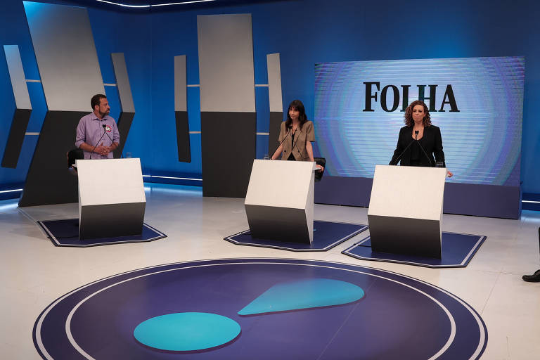 A imagem mostra um estúdio de debate com três participantes em pedestais. À esquerda, um homem com camisa roxa fala ao microfone. No centro, uma mulher com um vestido claro observa. À direita, uma mulher com cabelo cacheado e um casaco escuro também está em pé. Ao fundo, há um telão com a palavra 'FOLHA'. O chão do estúdio é circular com um design gráfico em azul.