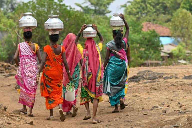 A imagem mostra um grupo de cinco mulheres caminhando em uma estrada rural. Elas estão vestidas com saris coloridos e carregam potes de metal na cabeça. Algumas mulheres também têm baldes ou recipientes nas mãos. Ao fundo, há vegetação e algumas casas visíveis.
