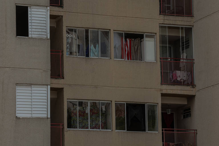 A imagem mostra a fachada de um edifício residencial de cor bege. Várias janelas estão visíveis, algumas com cortinas e roupas penduradas. Há varandas com grades em algumas janelas. A iluminação é suave, sugerindo um dia nublado.