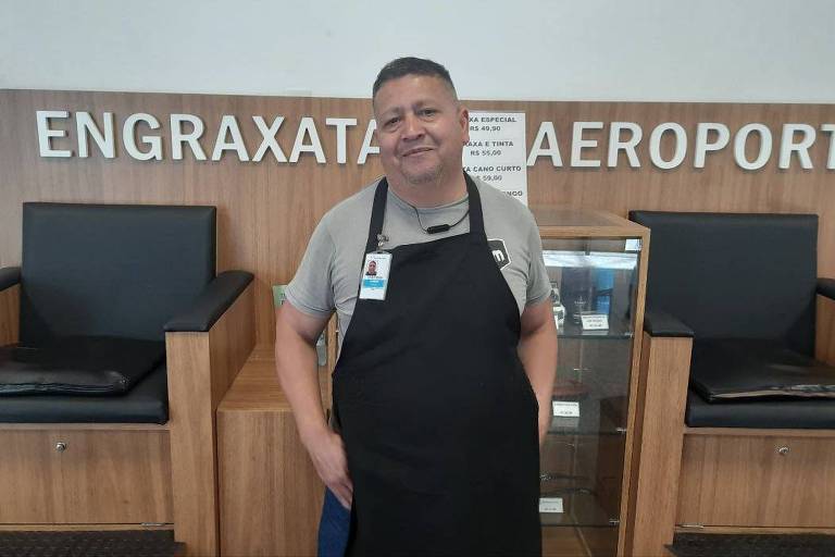  Um homem está posando em frente a um balcão de engraxate no aeroporto. Ele usa uma camiseta cinza e um avental preto. Ao fundo, há uma placa com a palavra 'ENGRAXATA AEROPORTO' e uma vitrine com produtos. Dois cadeirões pretos estão visíveis ao lado.