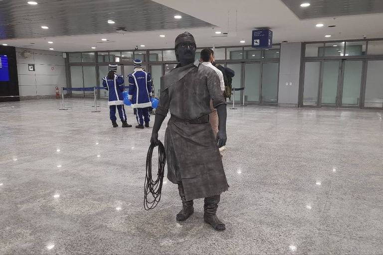  A imagem mostra um artista de rua pintado de cinza, vestido com roupas que lembram um personagem de época, segurando um laço. Ele está posicionado em um aeroporto, com painéis de informações de voos ao fundo. Há outras pessoas ao redor, algumas com malas, e um ambiente amplo e moderno.