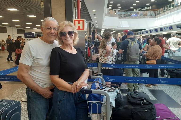  Um casal sorridente posa para a foto em um aeroporto. O homem usa uma camiseta branca e óculos, enquanto a mulher veste uma blusa preta e também usa óculos escuros. Eles estão em frente a um carrinho de bagagem, com várias malas ao fundo. O ambiente está movimentado, com muitas pessoas na fila de check-in e placas indicando 'Porto Alegre'.