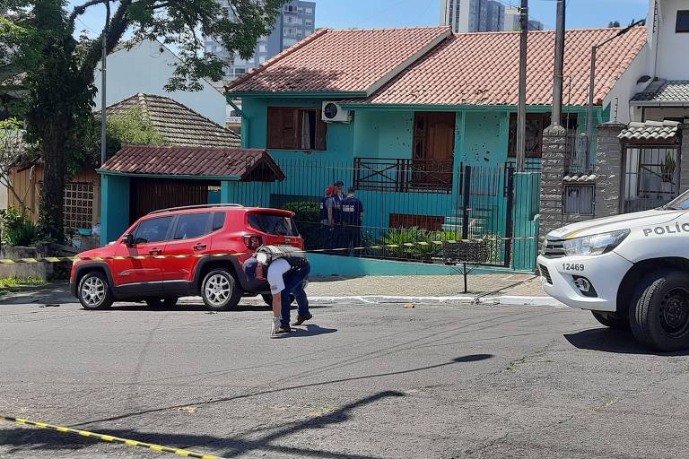 A imagem mostra uma rua em um ambiente urbano. À esquerda, há um carro vermelho estacionado e, à direita, um veículo da polícia. No fundo, uma casa de cor azul com janelas e uma porta. Um policial está em pé próximo ao carro vermelho, enquanto uma fita de isolamento amarelo é visível no chão.