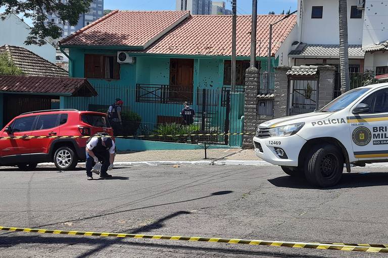 Na foto, um homem com farda policial recolhe projéteis de arma de uma via pública. Ao lado, uma caminhonete da brigada e um carro vermelho. Ao fundo, uma casa de paredes verdes, furadas de tiro.