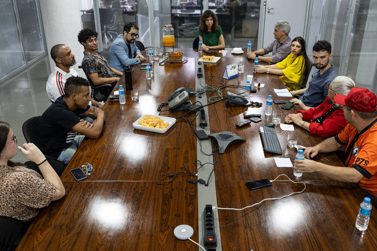 A imagem mostra uma reunião em uma mesa de conferência longa, com várias pessoas sentadas ao redor. Há um total de 12 pessoas visíveis, algumas usando roupas casuais e outras mais formais. No centro da mesa, há um prato com salgadinhos e garrafas de água. Ao fundo, é possível ver janelas de vidro e um ambiente de escritório. A iluminação é clara e o ambiente parece moderno.