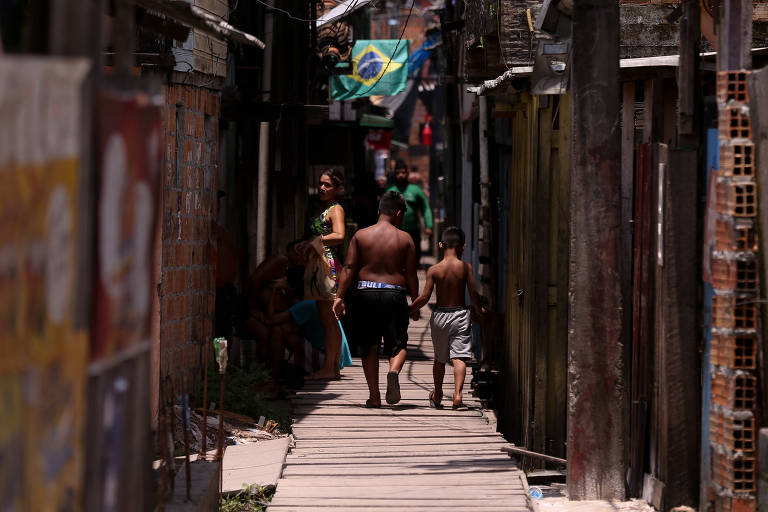 A imagem mostra uma rua estreita em uma comunidade, com casas de tijolos e madeira. Duas crianças, um menino e um garoto, caminham de mãos dadas. Ao fundo, uma mulher está sentada, enquanto outras pessoas estão visíveis nas laterais. Uma bandeira do Brasil está pendurada em um dos prédios.