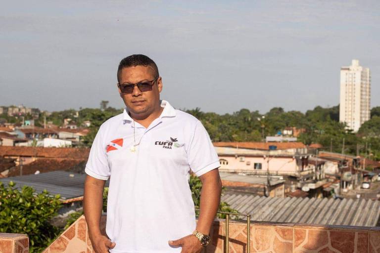 Um homem de óculos escuros e camisa branca está à frente de uma área de casas, com uma floresta e um prédio ao fundo