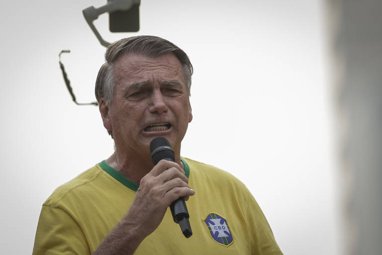 Um homem está falando ao microfone, vestindo uma camiseta amarela com detalhes verdes e um emblema do Brasil. Ele parece estar em um evento ao ar livre, com um fundo desfocado. O homem tem cabelo liso e está expressando emoções enquanto fala.